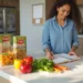 A woman stands in a kitchen, smiling as she writes in a notebook. On the counter are pasta boxes, a jar of pasta, colorful bell peppers, carrots, parsley, and a calculator to help with her grocery budget. | Gyyol.com