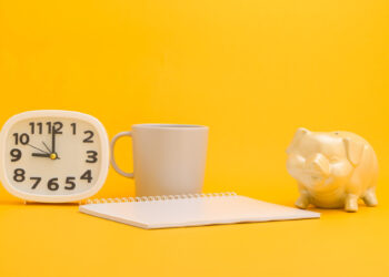 A white clock, a gray mug, a blank spiral notebook, and a white piggy bank are arranged on a bright yellow background—perfect for planning your short term savings goals. | Gyyol.com