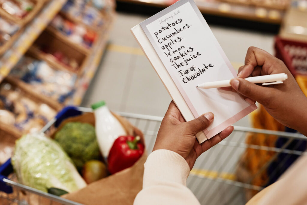 A person holding a shopping list and pen above a grocery cart filled with produce, milk, and a red bell pepper in a supermarket aisle, carefully sticking to their grocery budget while checking off potatoes, cucumber, apples, lemon, cookies, and more. | Gyyol.com