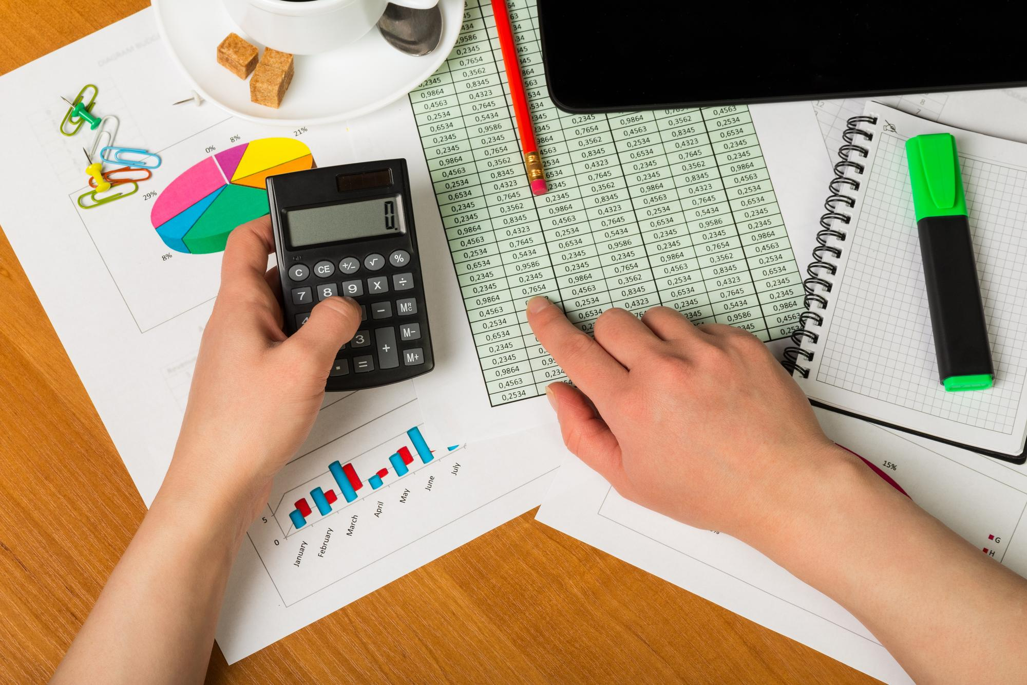 A person uses a calculator and points to a monthly income figure on a printed spreadsheet, surrounded by charts, graphs, a notebook, highlighters, and a cup of coffee on a wooden desk. | Gyyol.com