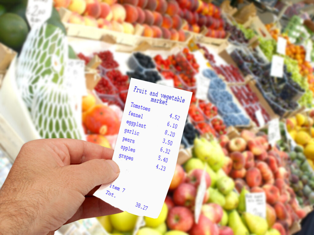 A hand holds a receipt listing fruits and vegetables with prices, highlighting a smart grocery budget, in front of a market stall overflowing with colorful fresh produce like apples, pears, grapes, and berries. | Gyyol.com
